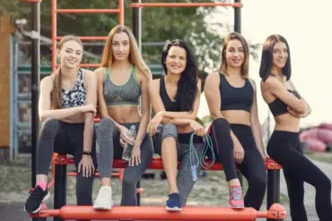 Five women in athletic wear sit and stand on outdoor gym equipment, smiling confidently. The setting is sunny and outdoors, conveying a sense of camaraderie and fitness.