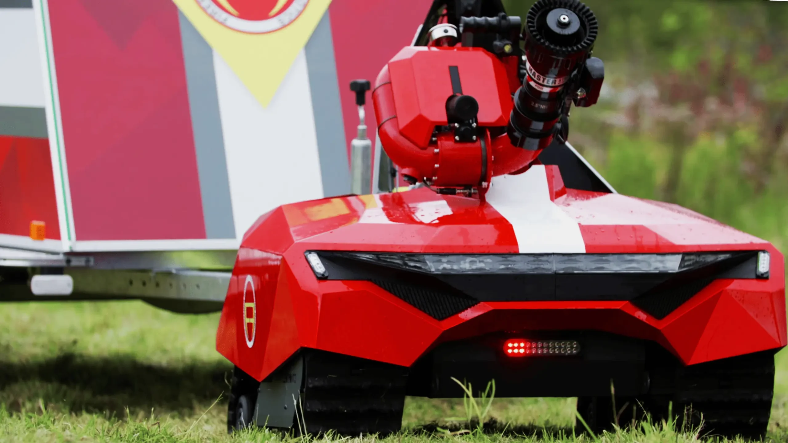 A red firefighting robot with tank-like treads and a mounted hose is on grass beside a vehicle. It appears ready for action, conveying efficiency.