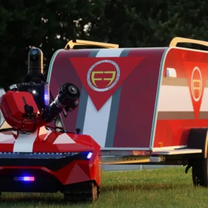 A futuristic red and white robotic vehicle on grass, featuring LED lights and a camera. It tows a matching trailer with a stylized logo, set against trees.
