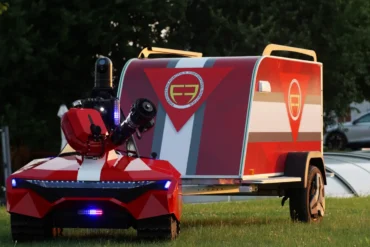 A futuristic red and white robotic vehicle on grass, featuring LED lights and a camera. It tows a matching trailer with a stylized logo, set against trees.