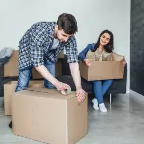 A couple packs boxes in a bright living room. The man tapes a box, while the woman sits on the couch smiling, holding a smaller box. The mood is cheerful.