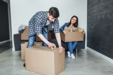A couple packs boxes in a bright living room. The man tapes a box, while the woman sits on the couch smiling, holding a smaller box. The mood is cheerful.