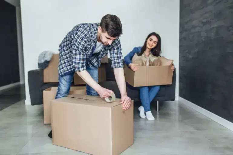 A couple packs boxes in a bright living room. The man tapes a box, while the woman sits on the couch smiling, holding a smaller box. The mood is cheerful.