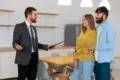 A real estate agent in a suit gestures while holding a clipboard, speaking to a smiling couple in a modern kitchen. The atmosphere is positive and engaging.