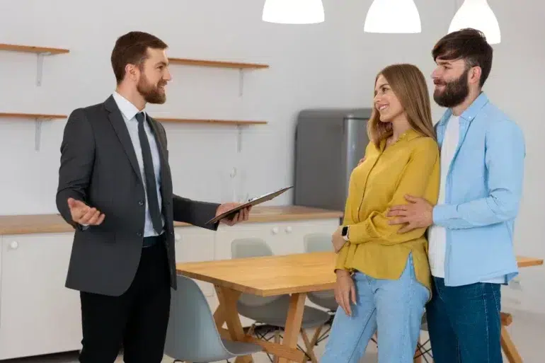 A real estate agent in a suit gestures while holding a clipboard, speaking to a smiling couple in a modern kitchen. The atmosphere is positive and engaging.