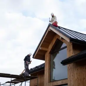 Two construction workers wearing helmets working on the metal roof of a modern wooden house under a cloudy sky.
