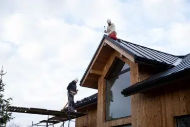 Two construction workers wearing helmets working on the metal roof of a modern wooden house under a cloudy sky.