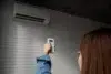 A person in a blue shirt points a remote control at a wall-mounted air conditioner on a textured white brick wall, adjusting settings.