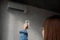 A person in a blue shirt points a remote control at a wall-mounted air conditioner on a textured white brick wall, adjusting settings.