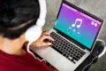 A person wearing white headphones is using a laptop with a colorful music player interface. The screen shows a large musical note, play buttons, and vibrant colors.