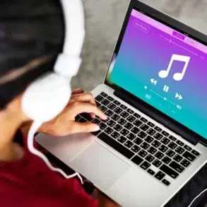 A person wearing white headphones is using a laptop with a colorful music player interface. The screen shows a large musical note, play buttons, and vibrant colors.