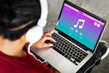 A person wearing white headphones is using a laptop with a colorful music player interface. The screen shows a large musical note, play buttons, and vibrant colors.