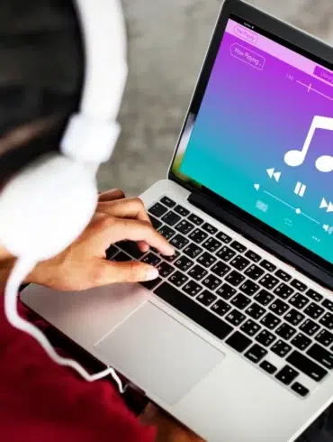 A person wearing white headphones is using a laptop with a colorful music player interface. The screen shows a large musical note, play buttons, and vibrant colors.