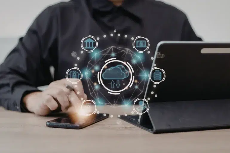Person using a smartphone and tablet with digital cloud computing and data network icons floating above the devices on a wooden table.