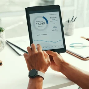 A person holds a tablet displaying financial graphs and data at a desk with notebooks, a phone, a plant, and a pen. The scene is focused and professional.
