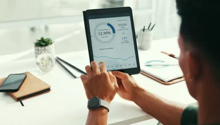 A person holds a tablet displaying financial graphs and data at a desk with notebooks, a phone, a plant, and a pen. The scene is focused and professional.