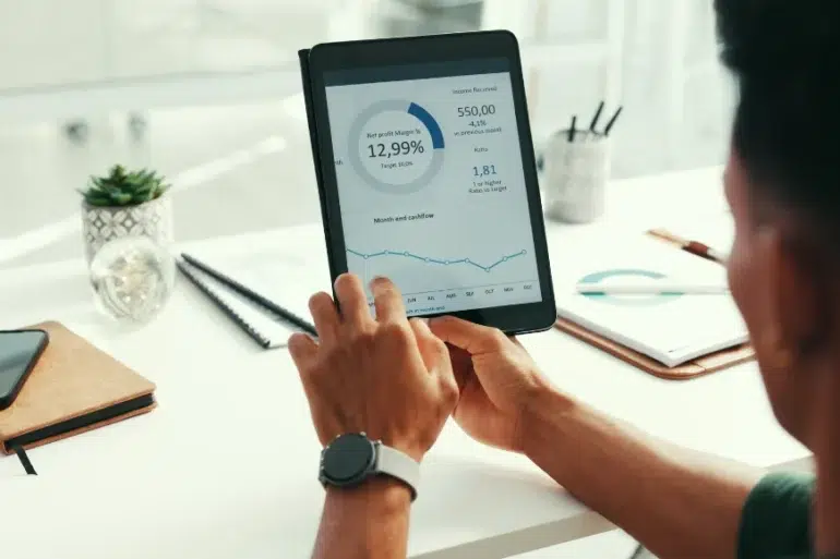 A person holds a tablet displaying financial graphs and data at a desk with notebooks, a phone, a plant, and a pen. The scene is focused and professional.