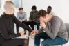 Therapist comforting a distressed man during a group counseling session with others seated in the background.