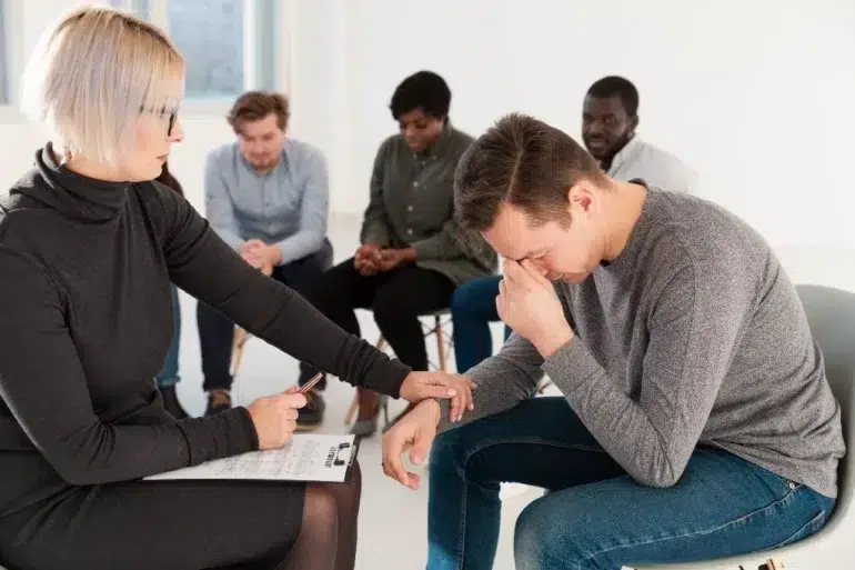 Therapist comforting a distressed man during a group counseling session with others seated in the background.
