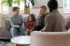 A family with a child sits on a sofa, talking to a counselor in a cozy, plant-filled room with soft lighting.