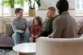 A family with a child sits on a sofa, talking to a counselor in a cozy, plant-filled room with soft lighting.