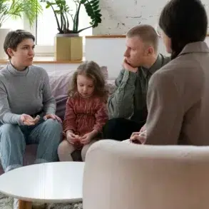 A family with a child sits on a sofa, talking to a counselor in a cozy, plant-filled room with soft lighting.