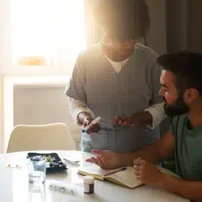 Healthcare professional explains medication to seated man at a sunlit table with medical supplies and notes.
