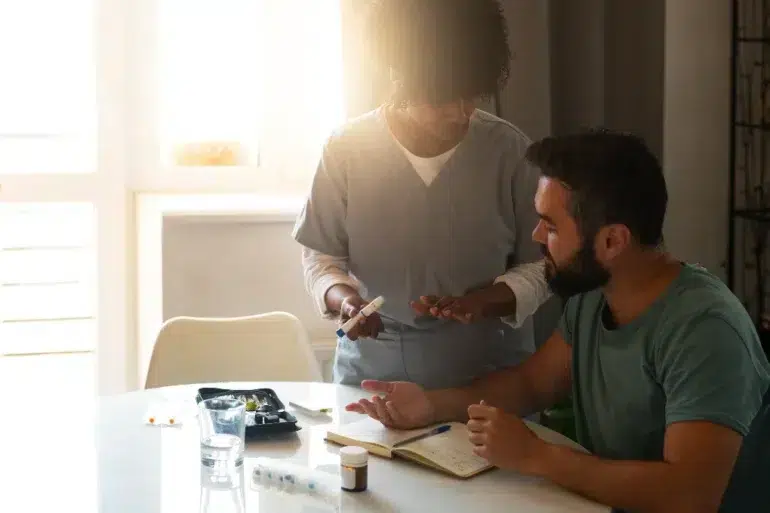 Healthcare professional explains medication to seated man at a sunlit table with medical supplies and notes.