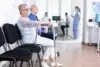 Elderly woman with a walker and a man sit in a clinic waiting room. A nurse and a patient are visible through glass doors. The scene is calm and clinical.