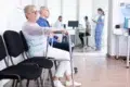 Elderly woman with a walker and a man sit in a clinic waiting room. A nurse and a patient are visible through glass doors. The scene is calm and clinical.