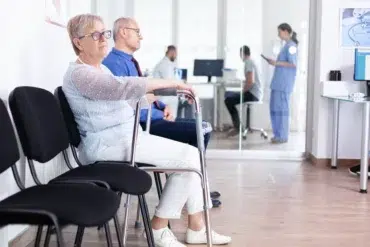 Elderly woman with a walker and a man sit in a clinic waiting room. A nurse and a patient are visible through glass doors. The scene is calm and clinical.