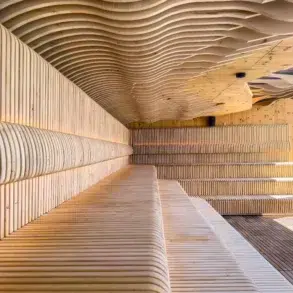 Interior of a modern wooden sauna with curved, slatted benches and ceiling designs, illuminated by warm lighting and natural daylight.