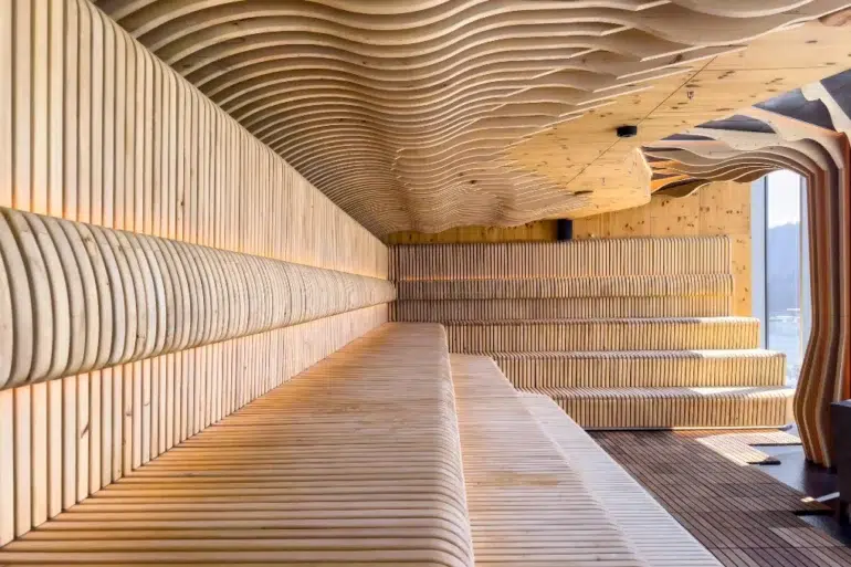 Interior of a modern wooden sauna with curved, slatted benches and ceiling designs, illuminated by warm lighting and natural daylight.