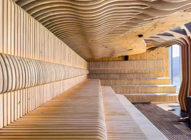 Interior of a modern wooden sauna with curved, slatted benches and ceiling designs, illuminated by warm lighting and natural daylight.