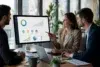 A group of business professionals in a modern office discuss performance metrics and reward tiers displayed on a large monitor, showcasing the capabilities of B2B Loyalty Program Software.