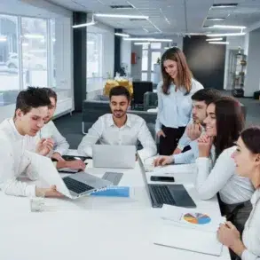 A group of seven young professionals in a modern office setting, collaborating around a table with laptops and documents, smiling and engaged.