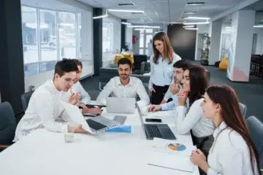A group of seven young professionals in a modern office setting, collaborating around a table with laptops and documents, smiling and engaged.
