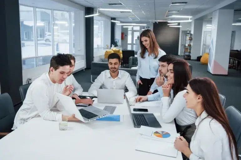 A group of seven young professionals in a modern office setting, collaborating around a table with laptops and documents, smiling and engaged.