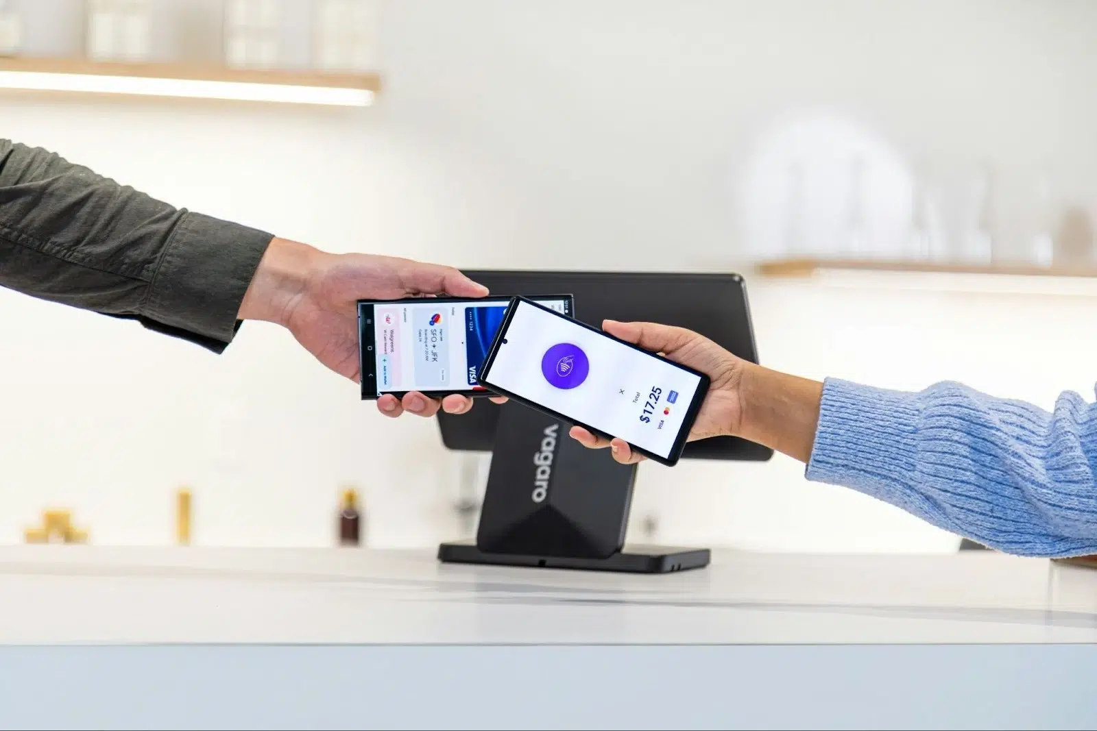 Two people exchange smartphones over a counter, displaying payment apps. A modern, cashless transaction scene. Background shows a sleek, minimal interior.