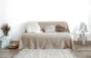 Minimalist living room with a beige sofa, assorted textured pillows, and a soft rug. A chair with a draped blanket adds warmth and coziness.