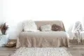 Minimalist living room with a beige sofa, assorted textured pillows, and a soft rug. A chair with a draped blanket adds warmth and coziness.