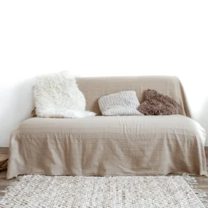 Minimalist living room with a beige sofa, assorted textured pillows, and a soft rug. A chair with a draped blanket adds warmth and coziness.