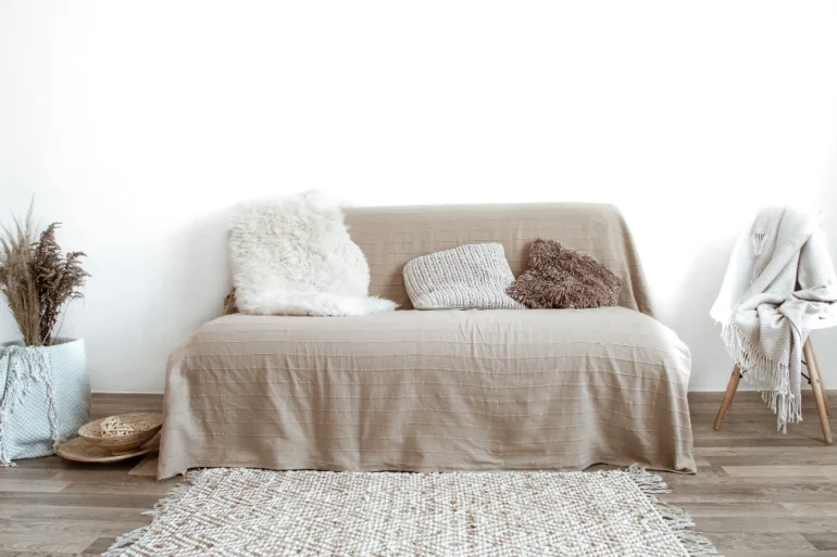 Minimalist living room with a beige sofa, assorted textured pillows, and a soft rug. A chair with a draped blanket adds warmth and coziness.