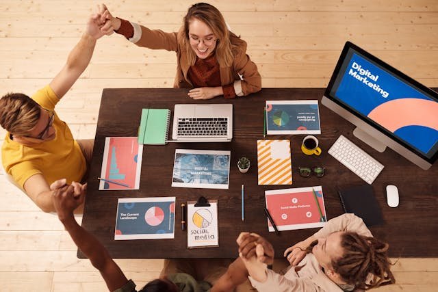 Overhead view of a diverse team celebrating around a desk with laptops and digital marketing materials, conveying teamwork and success.