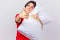 A woman in a red shirt hugs a white pillow and gives a thumbs up, smiling warmly. The background is light gray, conveying comfort and positivity.