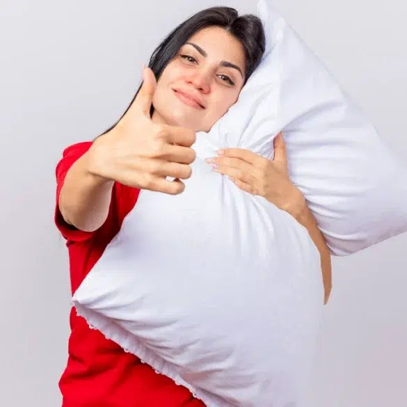 A woman in a red shirt hugs a white pillow and gives a thumbs up, smiling warmly. The background is light gray, conveying comfort and positivity.