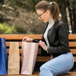 A woman in glasses and a black jacket sits on a park bench, smiling as she looks inside a shopping bag. Two more bags, blue and beige, sit beside her.