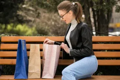 A woman in glasses and a black jacket sits on a park bench, smiling as she looks inside a shopping bag. Two more bags, blue and beige, sit beside her.