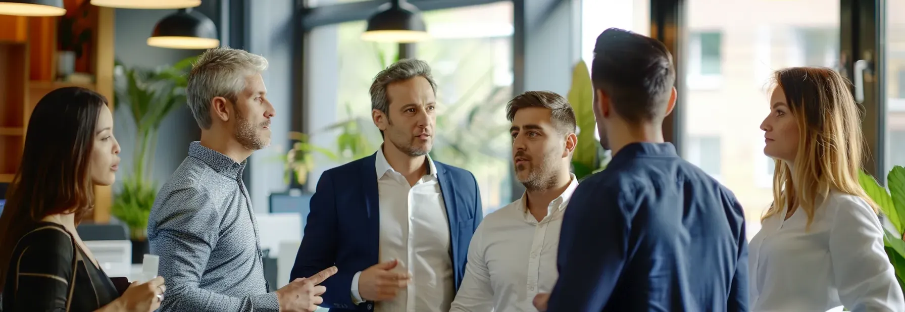A group of six professionals standing in a well-lit office, engaged in a serious discussion. The scene conveys collaboration and focus.