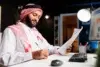 Man in traditional attire works at a desk, reviewing documents and jotting notes. A computer displaying charts is on, indicating a focused office setting.